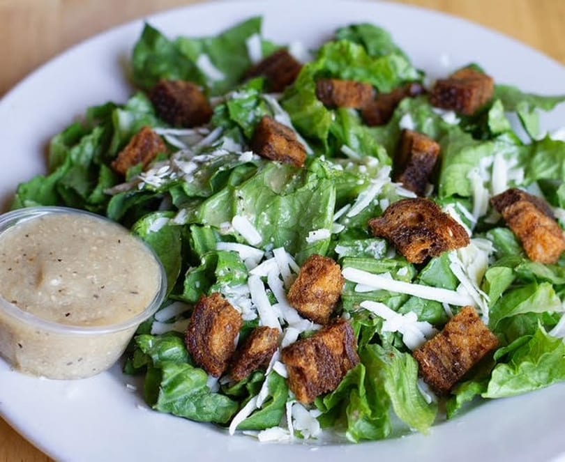 Caesar salad with green lettuce, shredded cheese, croutons, and a side of dressing.