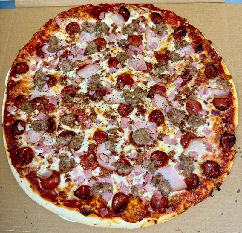 A whole meat lover's pizza with pepperoni, sausage, ham, and bacon in a cardboard box.